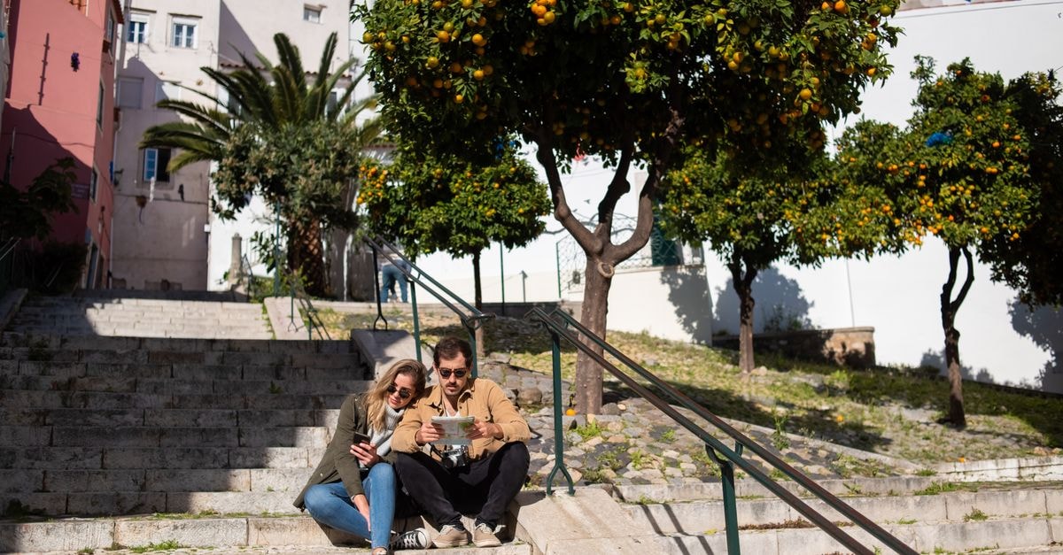 Leiria Travel Guide: History, City Style, and Local Culture Couple sitting on steps with a map, surrounded by vibrant orange trees in a sunny urban setting.