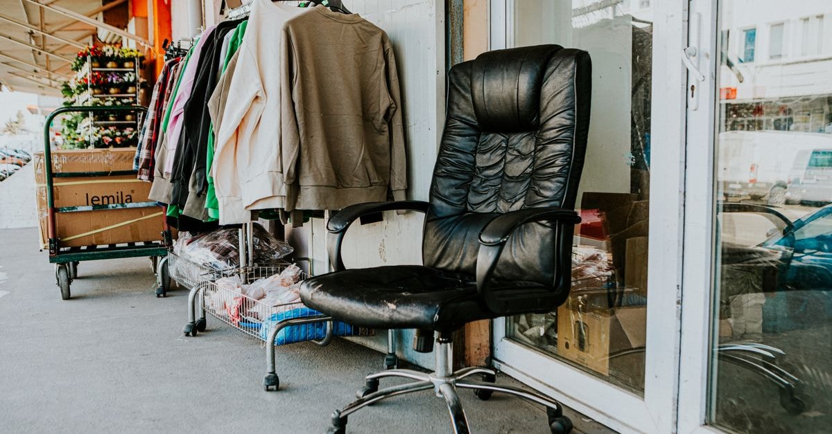 Worn black office chair outside a secondhand store with clothes and boxes nearby.