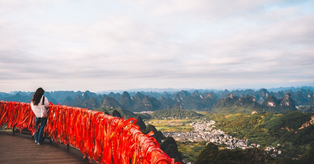 Qingyuan Travel Guide: River Light, Karst Peaks, and Slow Guangdong Days Traveler admiring stunning panorama from Fubo Hill, Guilin, with vibrant red ribbons and limestone mountains.
