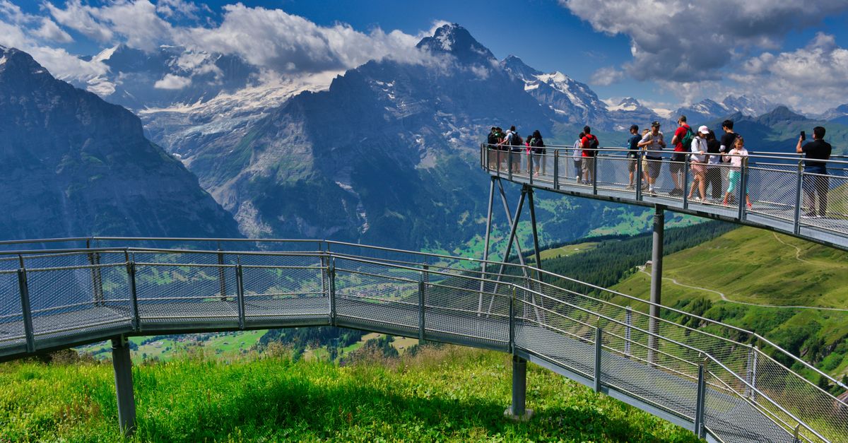 Bern in July 2026: A Practical Travel Guide for Peak-Summer Sightseeing Panoramic view of Grindelwald, Switzerland with tourists on a scenic bridge against majestic mountains.