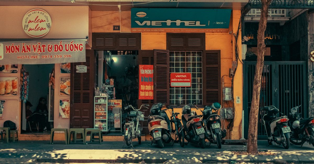 Colorful urban street scene with parked scooters, shops, and tree shadows in daylight.