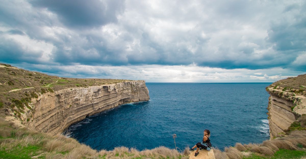A Quiet Maltese Seaside: A Birżebbuġa Travel Guide Woman seated on a cliff overlooking a dramatic coastal landscape under cloudy skies.
