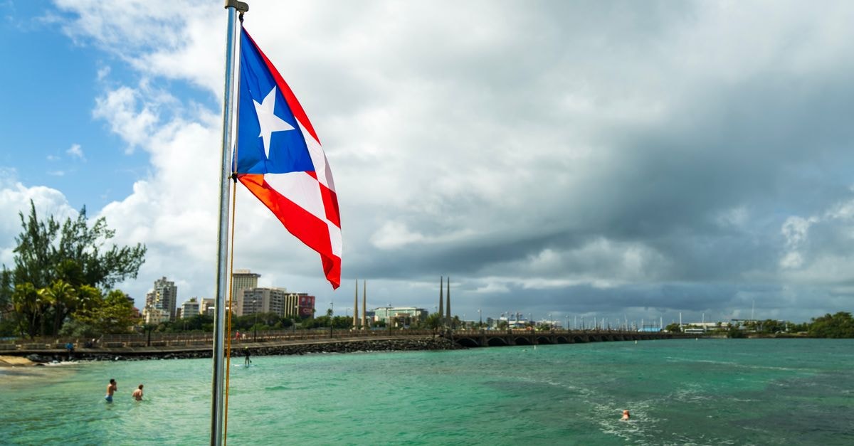 Hotel Las Caobas, San Francisco de Macorís — Pool, Tennis and a Quiet Avenida Setting Puerto Rican flag on a flagpole by the sea with city skyline in the background.