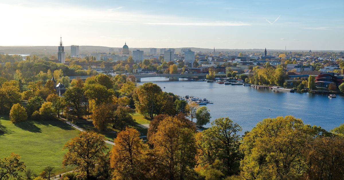 Aerial view of a lush park by a river with a city skyline in the background during autumn.