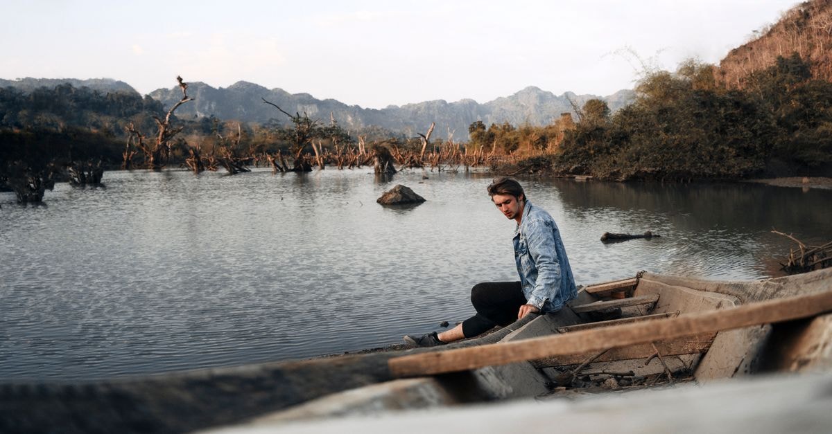 A man in a denim jacket sits in a boat on a serene Laos river with a mountainous backdrop.