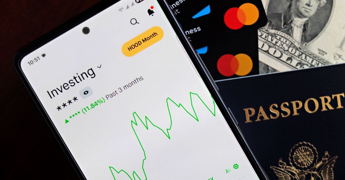 MetaMask and Mastercard launch self-custody crypto card in the U.S. Smartphone displaying investing app, with credit cards, cash, and passport nearby, symbolizing finance