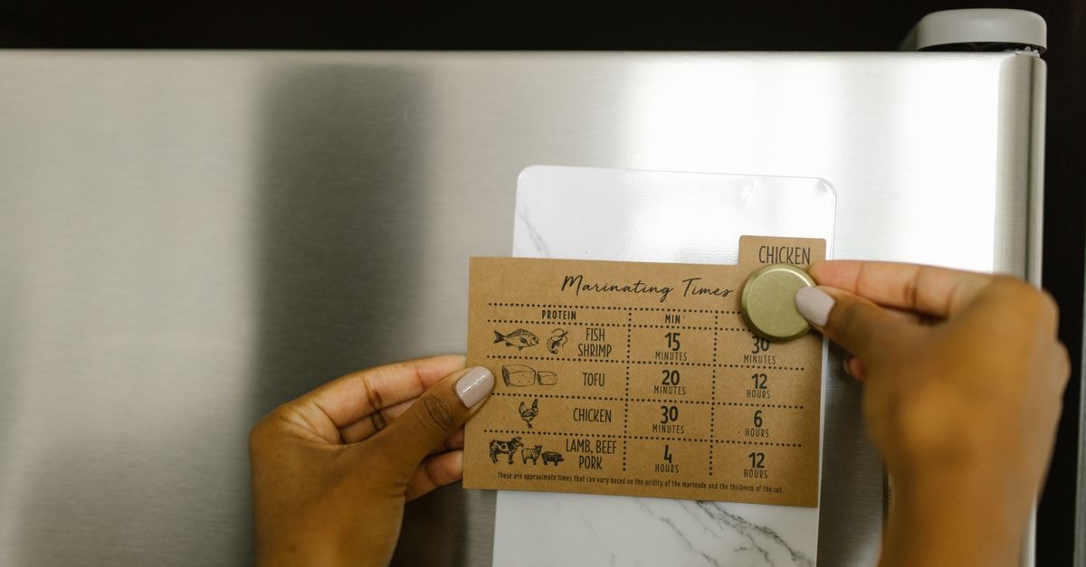 Hands placing a marinating timer card on a fridge with a magnet, featuring cooking times for various proteins.