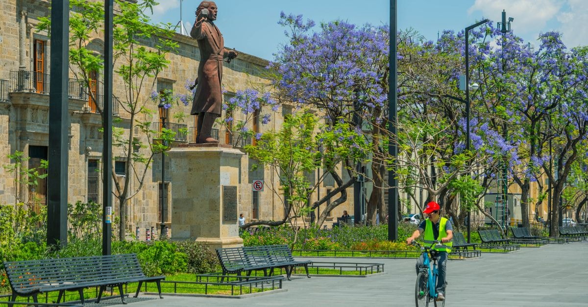 Guadalajara Travel Guide: Things to Know Before You Visit Cyclist rides by historic monument and jacaranda trees in a sunny Guadalajara park.