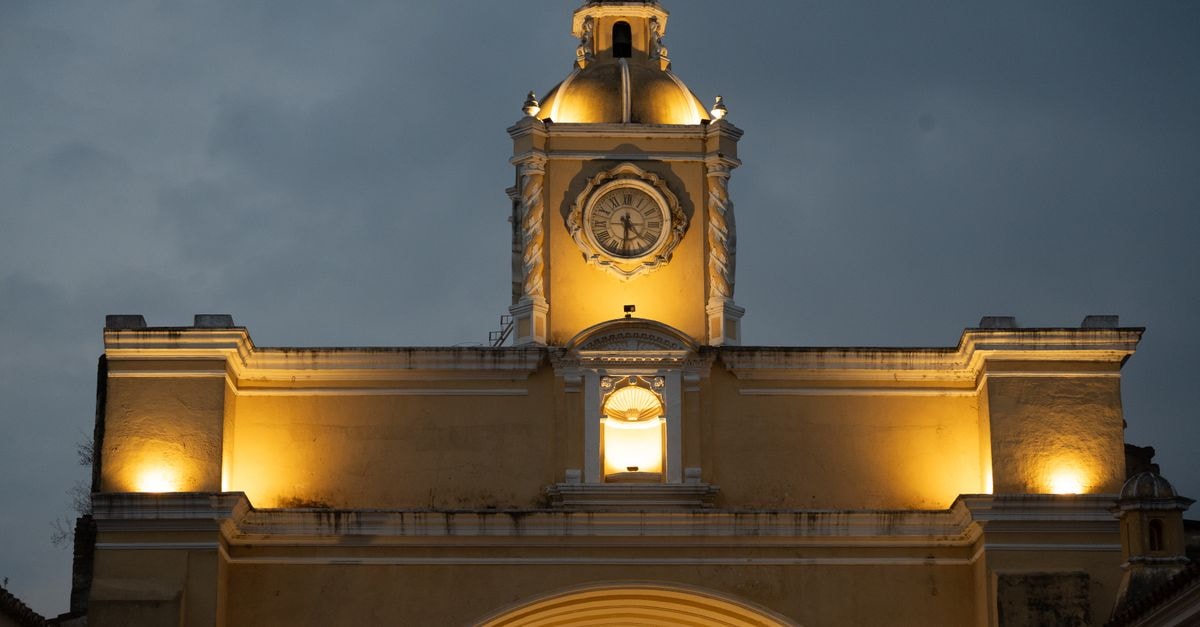 The illuminated Arco de Santa Catalina in Antigua, Guatemala, seen during the dusk against a cloudy sky.