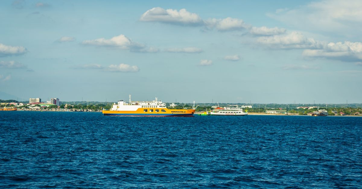 A vibrant yellow ferry sails across a deep blue sea with a coastal town in the background under a blue sky.