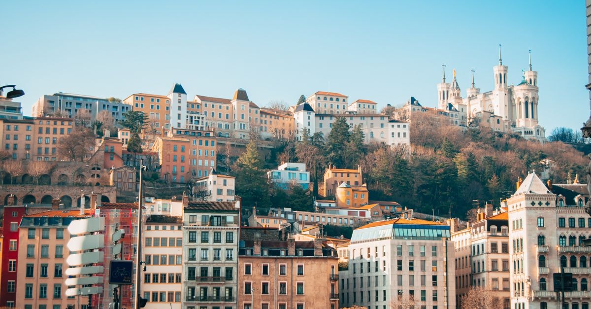 A vibrant cityscape of Lyon featuring the Basilica, buildings, and lush greenery on a hill.