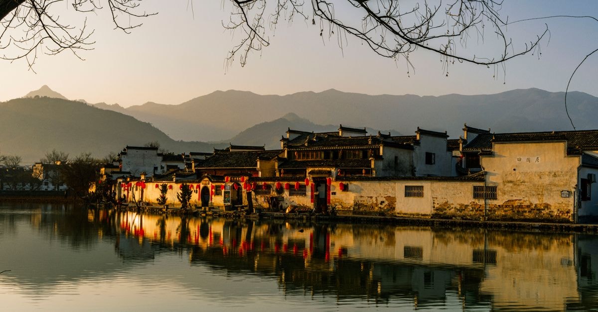 Anqiu Travel Guide: Ancient Roots and Rustic Landscapes A tranquil landscape of an Asian village with calm waters and mountain backdrop, captured at sunset.