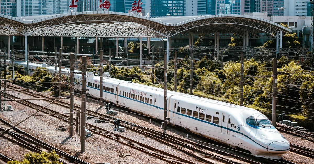 A high-speed train passing through Shanghai Railway Station with city skyline in the background.