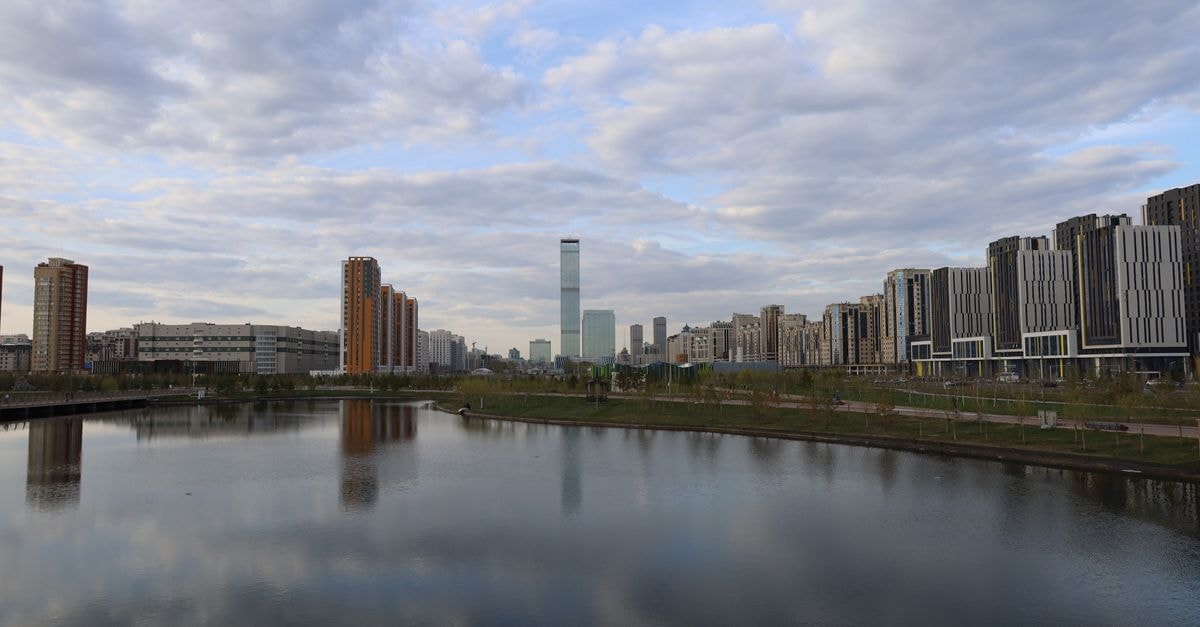 A modern city skyline reflected in a calm lake under a cloudy sky.