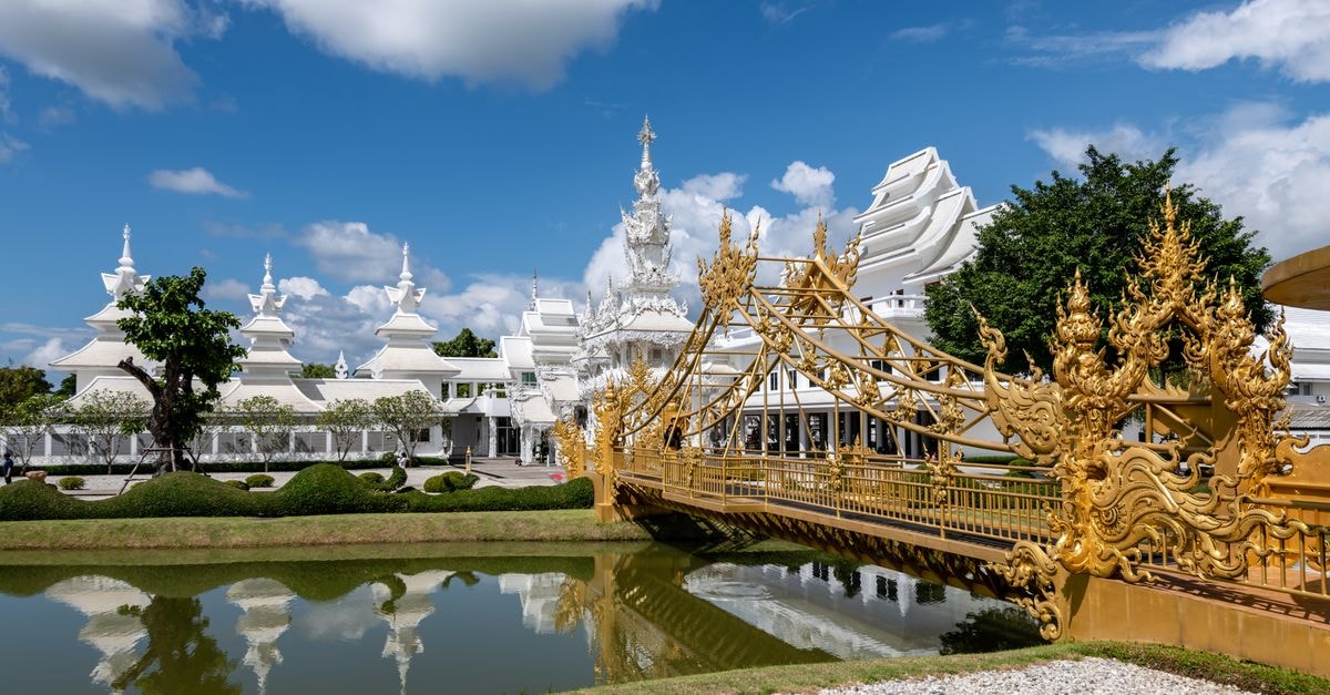 Golden bridge and Wat Rong Khun in Thailand, reflecting in a serene canal on a sunny day.
