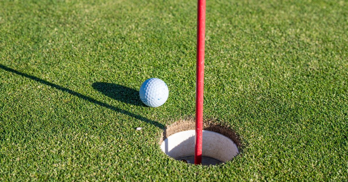 A close-up of a golf ball near a hole on a green with a red flagpole, symbolizing precision and sport.