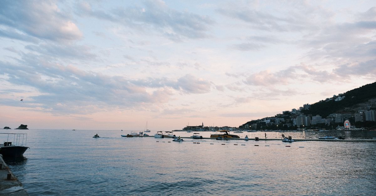 Serene sunset over Budva Bay with scattered boats and distant city skyline. Calm sea and pastel sky create a tranquil scene.