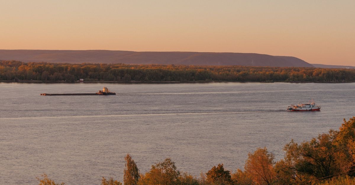 A tranquil river scene at sunset featuring two boats sailing with hills and autumn trees in the background.