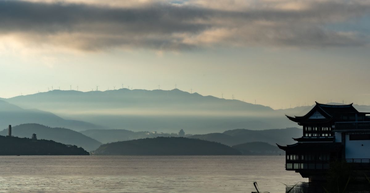 Peaceful sunrise over a serene lake with a traditional pagoda and misty hills in the background.