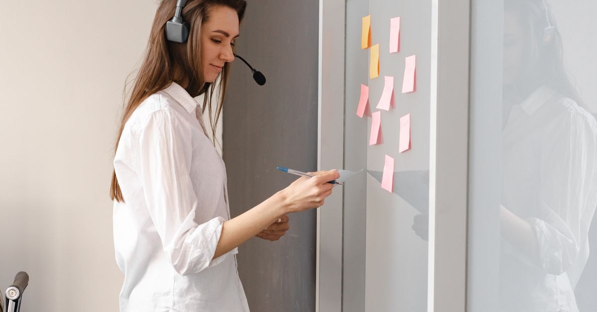 TaskUs: Levered Outsourcer at a Cyclical Trough Woman wearing headset organizing tasks on a glass board with sticky notes in an office setting.