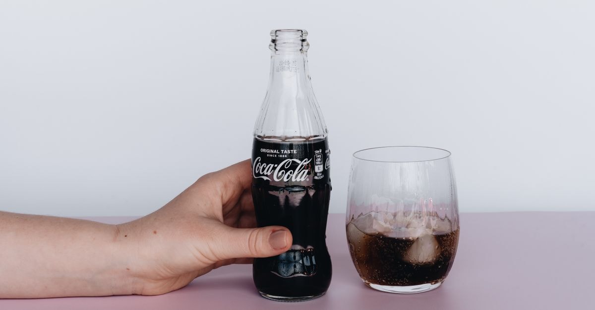 Close-up of a hand holding a Coca-Cola bottle next to a glass filled with ice and soda.