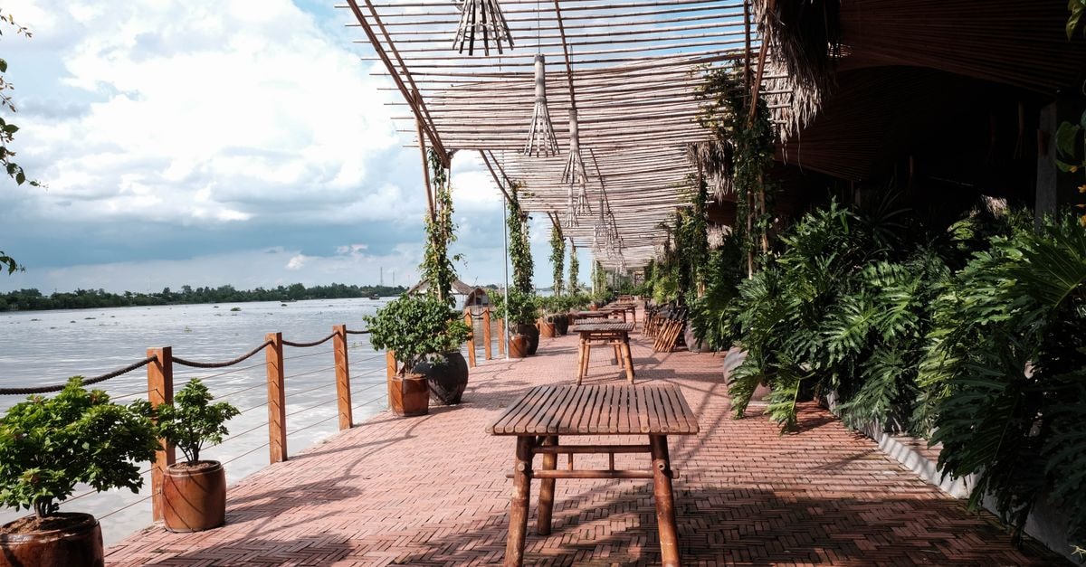Sunny day at a riverside terrace with wooden tables and lush plants, perfect for relaxation.