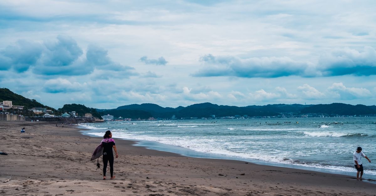People strolling along Kamakura beach, Japan on a cloudy day, with scenic ocean waves and distant hills.