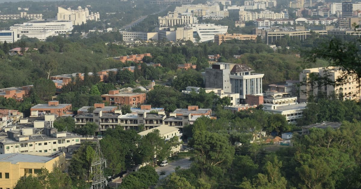 Overhead view of a cityscape surrounded by greenery under a clear sky, emphasizing urban architecture.