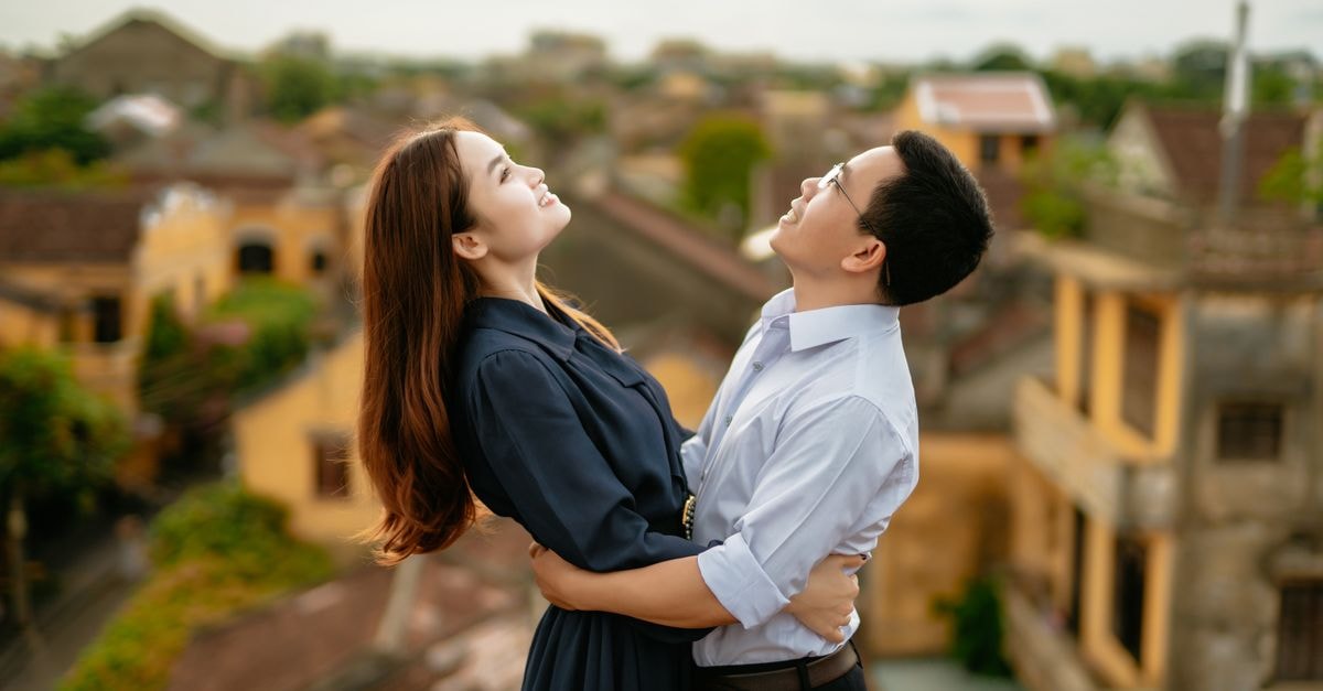 Side view of happy ethnic couple in elegant clothes embracing on balcony against town in summer day under gray sky while looking up