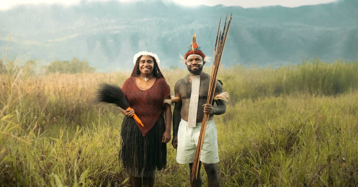 Couple in traditional dress stands in lush grassland with mountainous background.