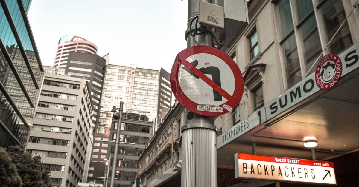Urban cityscape featuring a no U-turn sign on Queen Street, Selandia Baru, with modern high-rise buildings.